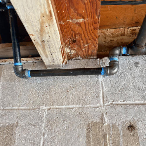 Black iron gas pipe with a vertical offset rising into wooden floor joists to feed an appliance on the floor above.