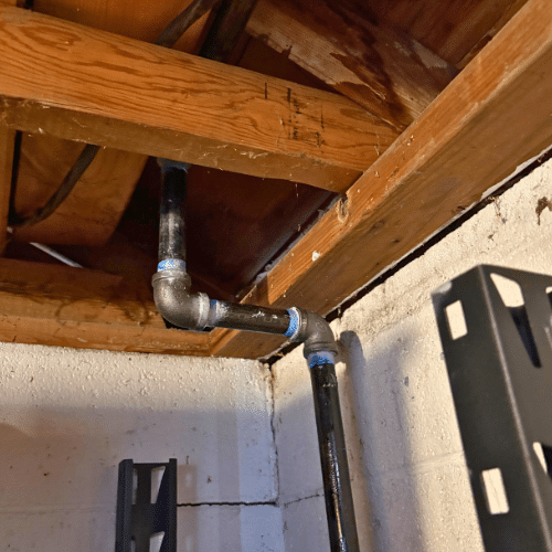Black iron gas pipe with a vertical offset rising into wooden floor joists to feed an appliance on the floor above.