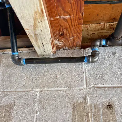 Black iron gas pipe with a vertical offset rising into wooden floor joists to feed an appliance on the floor above.