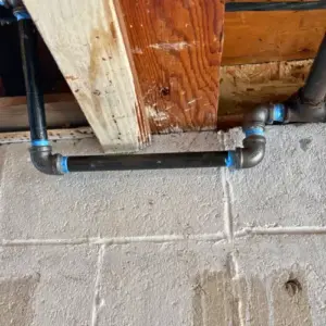 Black iron gas pipe with a vertical offset rising into wooden floor joists to feed an appliance on the floor above.