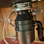 New black garbage disposal locked under the sink with mounting ring, drain elbow, and supply lines in a Midvale condo cabinet.