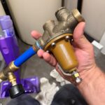 Technician holding removed brass builder-grade pressure reducing valve after diagnosis in North Salt Lake home