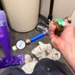 Hand holding old PRV body with separate downstream pressure gauge still attached on short blue PEX stub