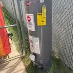 Old gray Rheem Performance Plus tank removed outdoors beside fence after North Salt Lake water heater replacement.