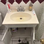 Open vanity cabinet with bathroom sink faucet work underway and new fixture parts staged on the floor