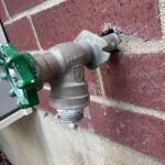 Old outdoor frost-free hose bib with green handle on red brick exterior wall before replacement in Sandy Utah