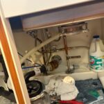Open under-sink cabinet showing disposal, copper lines, tools, and bleach during service